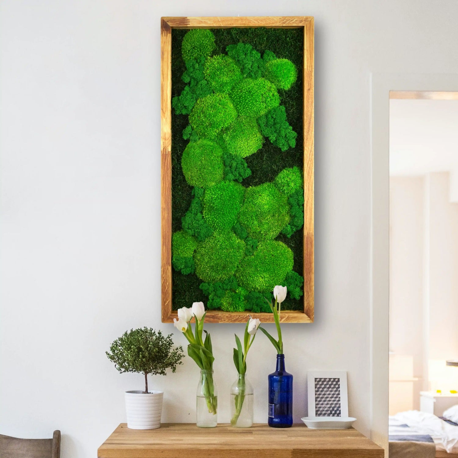 Handgemachtes Moosbild „Growth“ in einem Holzrahmen - minimalistische Wanddekoration aus grünen Moosen an einer weißen Wand in einem hellen, modern eingerichteten Raum. 